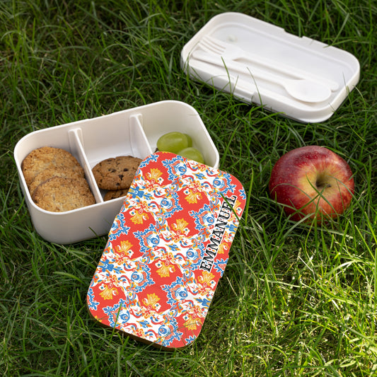 A colorful, African-inspired lunch box with a red, floral-patterned lid labeled EMMANUELLE sits open on grass. Inside, three sections hold cookies, a chocolate chip cookie, and green grapes. Nearby are a red apple and BPA-free container with cutlery.