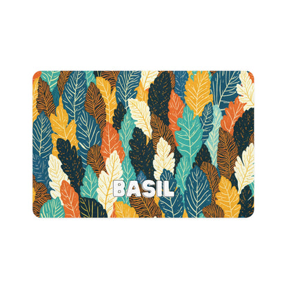 A rectangular card with an African-inspired pattern of overlapping leaves in teal, orange, yellow, white, and dark green. The word BASIL is centered at the bottom in bold, white, uppercase letters with a shadow effect on a white background.
