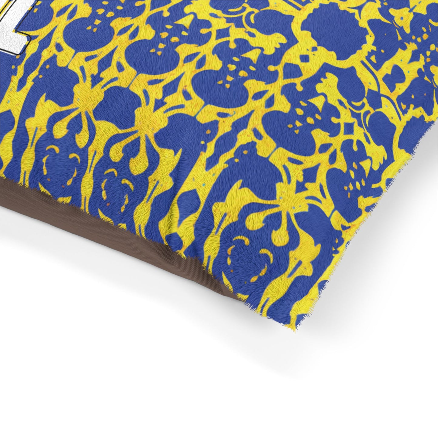 Close-up of a pillow with textured fabric. The pillow features an African-inspired blue and yellow ornate, floral-like pattern and white embroidered text partially visible. The background behind the pillow is plain and light-colored.