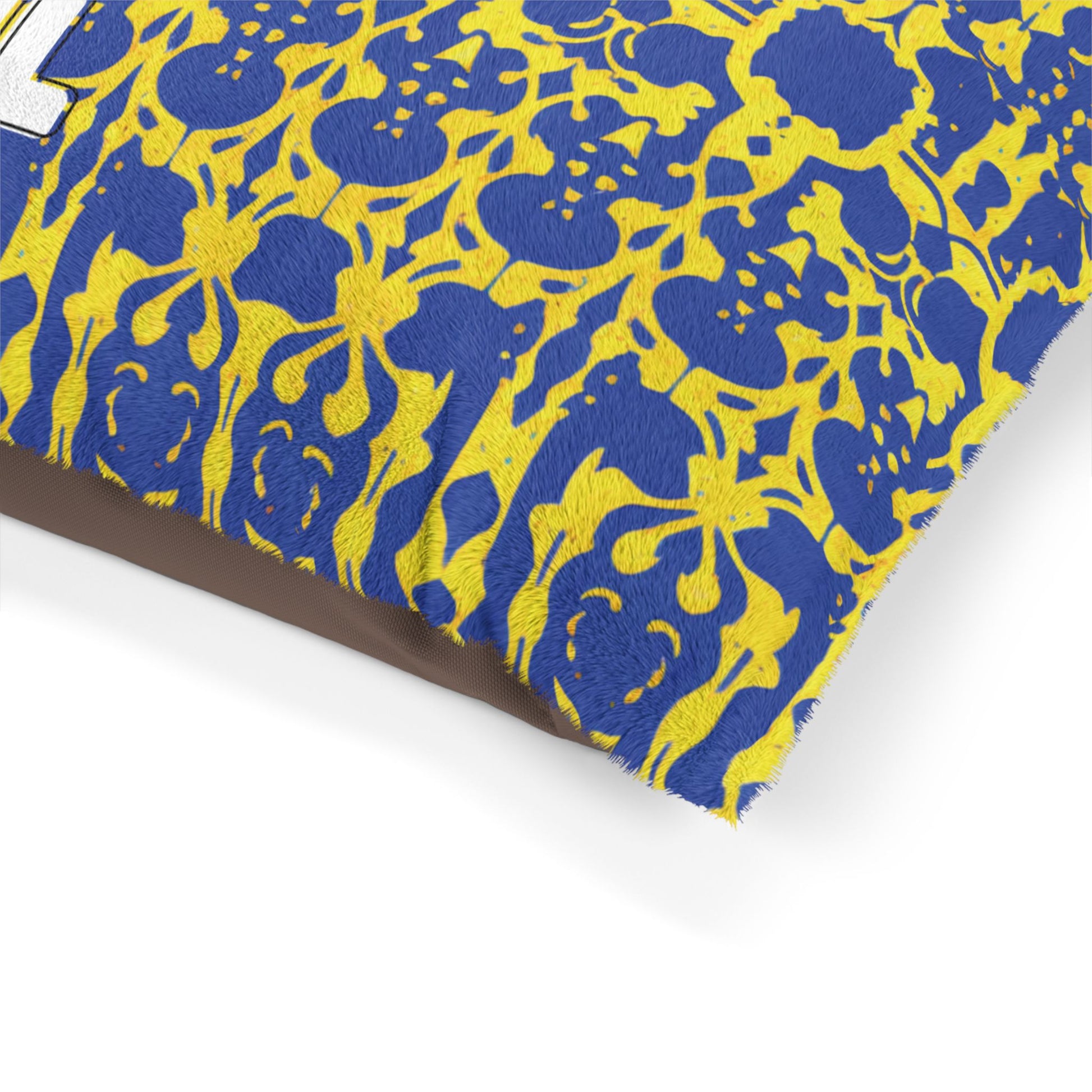 Close-up of a pillow with textured fabric. The pillow features an African-inspired blue and yellow ornate, floral-like pattern and white embroidered text partially visible. The background behind the pillow is plain and light-colored.