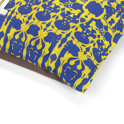 Close-up of a pillow with textured fabric. The pillow features an African-inspired blue and yellow ornate, floral-like pattern and white embroidered text partially visible. The background behind the pillow is plain and light-colored.