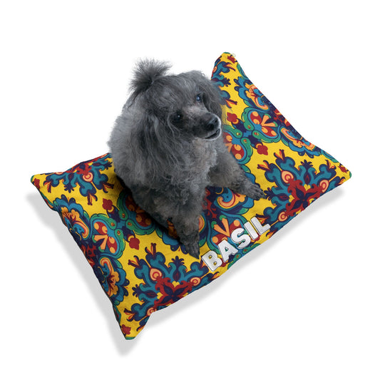 A gray poodle with curly fur sits on a cozy dog bed featuring a vibrant yellow, red, and blue floral pattern. The pillow, personalized with the name BASIL in bold white letters on one corner, adds charm against the white background.