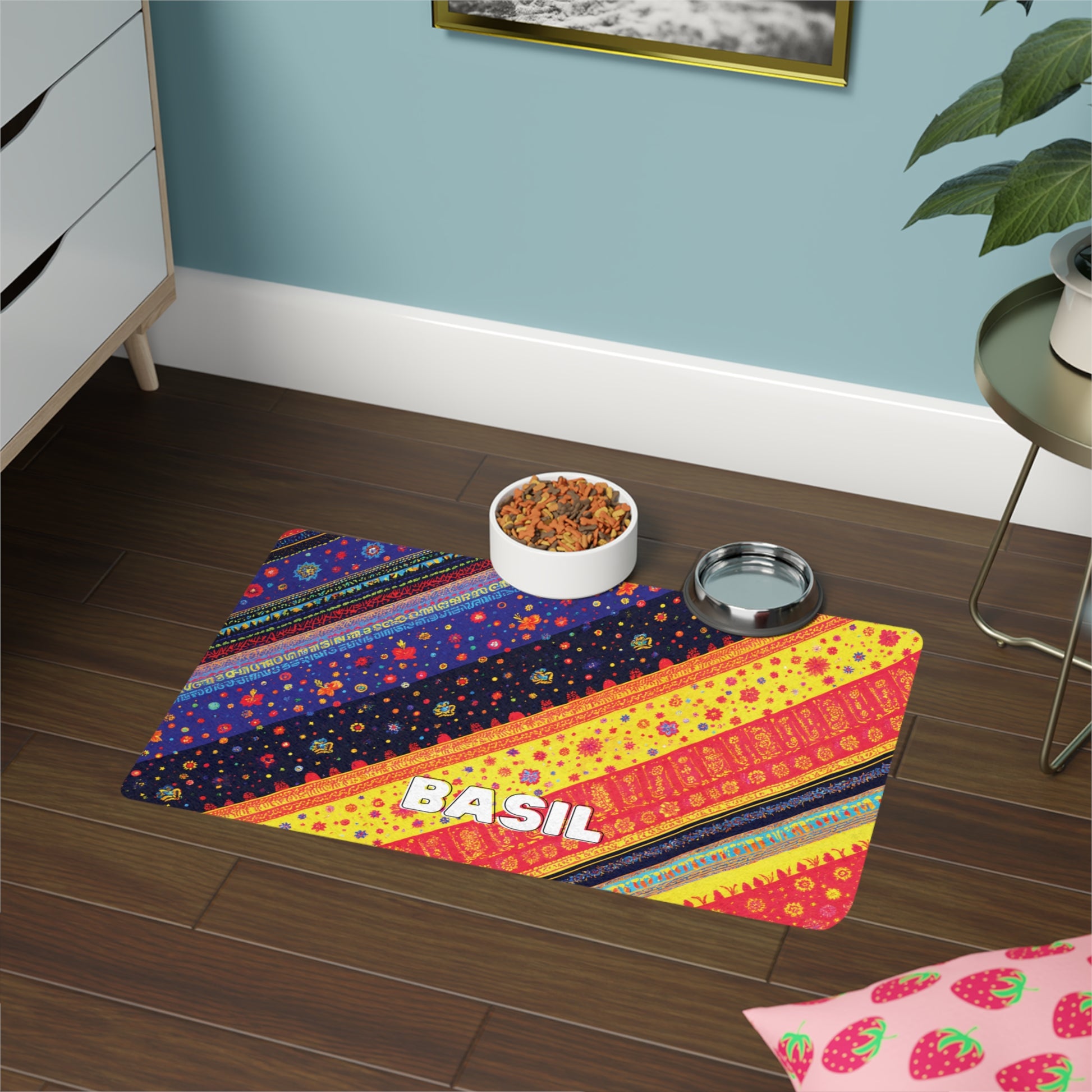 A colorful, African-inspired pet food mat with the name BASIL in bold white letters sits on a dark wooden floor. The mat’s vibrant patterns in yellow, red, and blue brighten the space near a plant, furniture, and strawberry-patterned pillow.