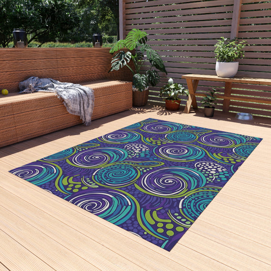 A modern outdoor patio with light wooden decking features an African-inspired outdoor rug with swirling blue, turquoise, and purple patterns. A wicker sofa with gray blanket, wooden table with plants, and lush greenery complete the cozy, sunlit space.