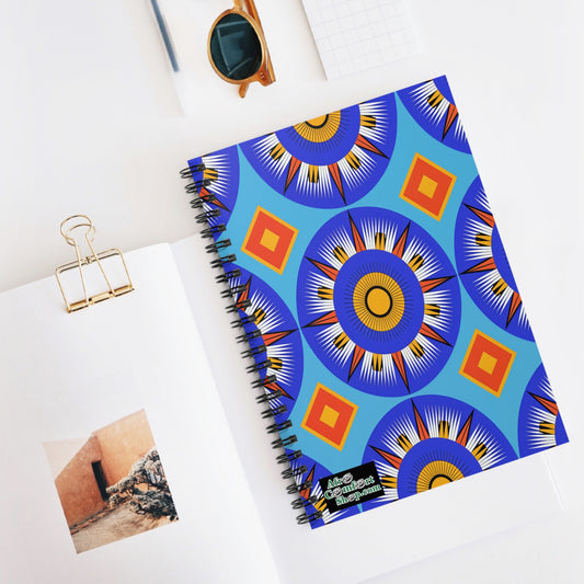 An African-inspired spiral notebook with a vibrant blue, yellow, red, and black geometric sunburst pattern lies on a white surface, partially covering an open notebook displaying a photo of a rustic building. Nearby are sunglasses and a gold binder clip.