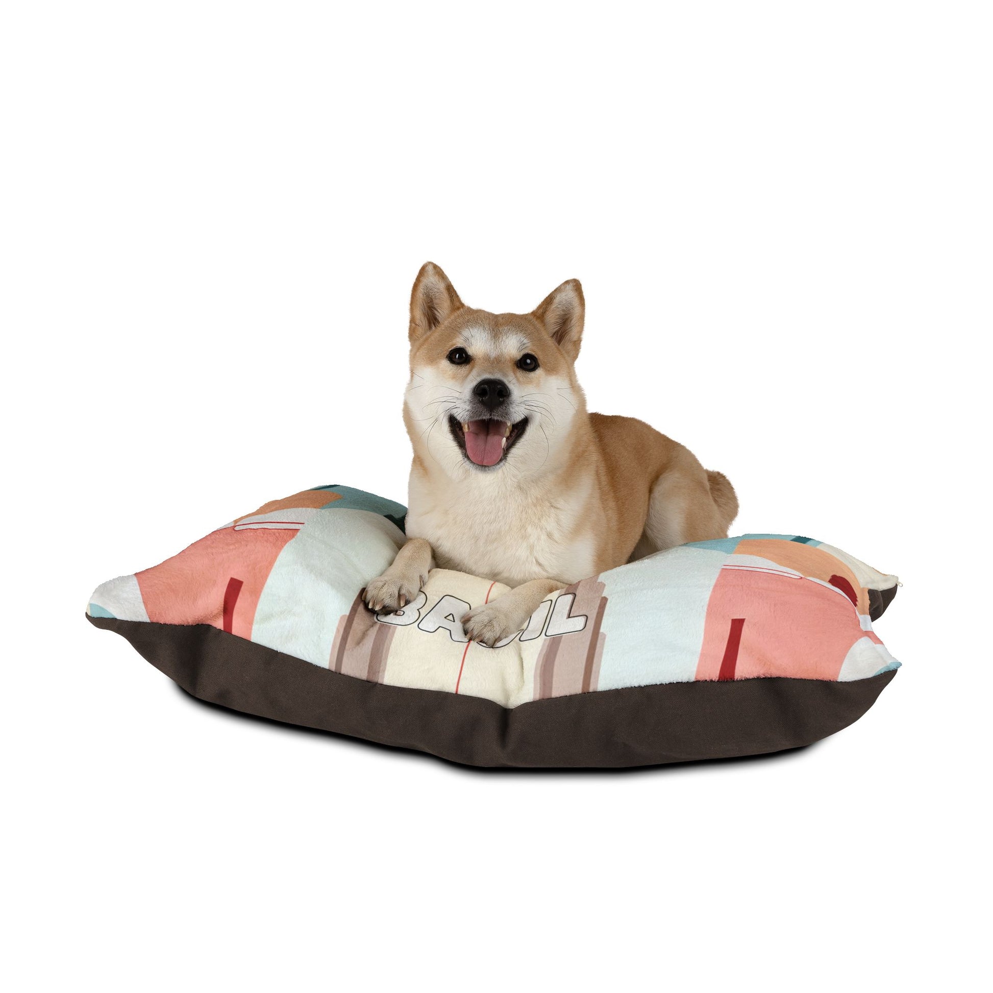 A happy Shiba Inu dog with tan fur lies on a colorful, African-inspired rectangular pillow, looking at the camera with its mouth open and tongue out. The pillow features pastel colors and geometric patterns against a plain white background.