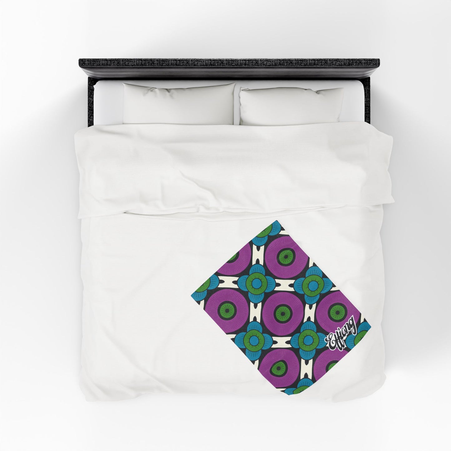 A neatly made bed with white bedding viewed from above. A folded, colorful African-inspired personalized blanket with a geometric pattern in blue, green, and purple lies diagonally at the foot, featuring the word energy in white script on one corner.