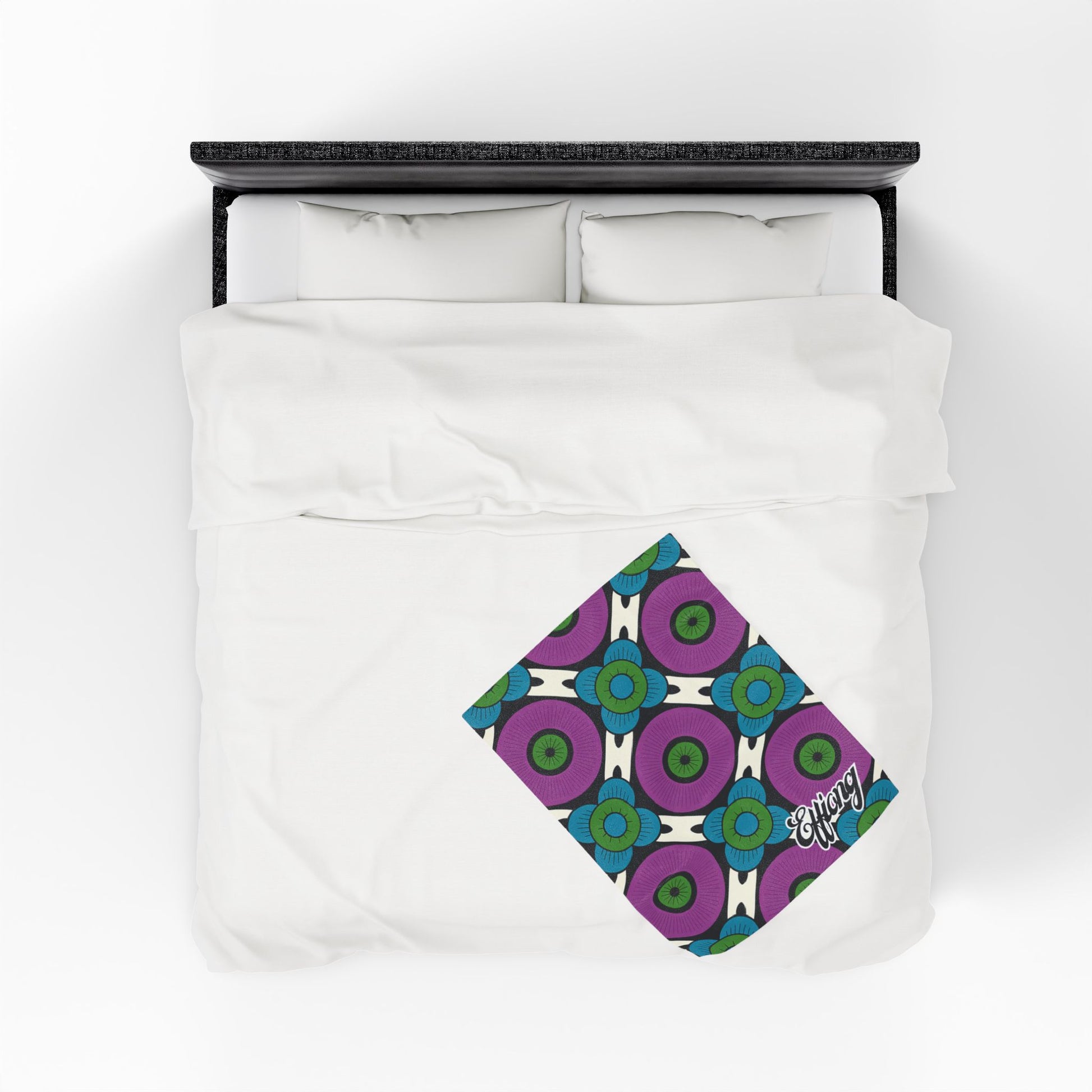 A neatly made bed with white bedding viewed from above. A folded, colorful African-inspired personalized blanket with a geometric pattern in blue, green, and purple lies diagonally at the foot, featuring the word energy in white script on one corner.
