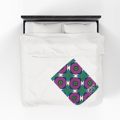 A neatly made bed with white bedding viewed from above. A folded, colorful African-inspired personalized blanket with a geometric pattern in blue, green, and purple lies diagonally at the foot, featuring the word energy in white script on one corner.
