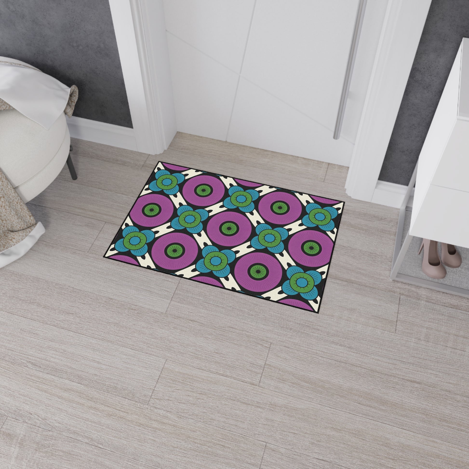 A geometric-patterned, non-slip doormat with purple, blue, green, and white circular designs lies on light wood flooring near a white cabinet and ottoman in a modern, gray-walled entryway. Shoes are visible on the bottom shelf of the cabinet.