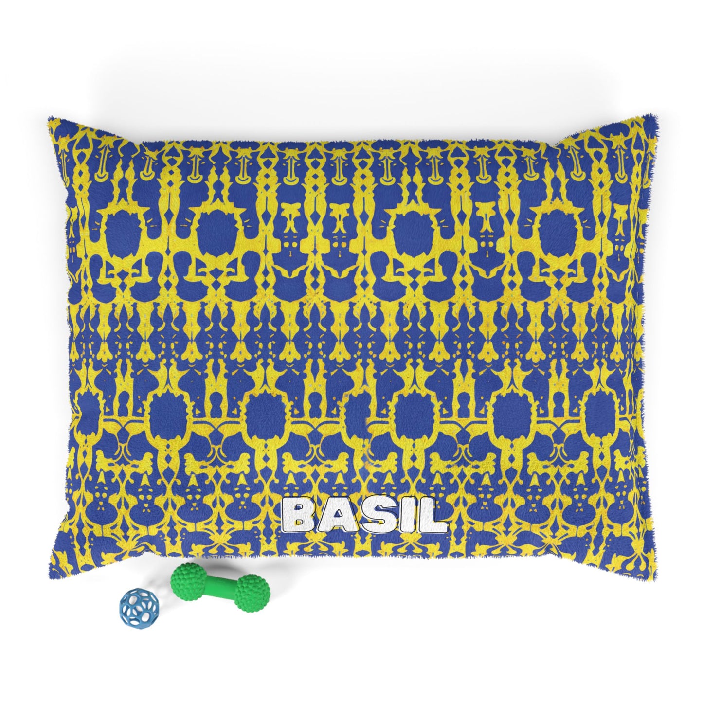 A rectangular cushion featuring a blue and yellow geometric pattern and “BASIL” in bold white letters offers luxurious comfort. Two green dog toys and a blue ball sit beside this personalized pet bed on a white background.
