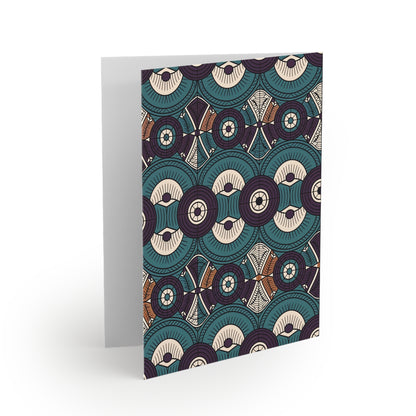 A greeting card stands upright, featuring an intricate pattern of overlapping teal, cream, and brown circular and geometric shapes with fine lines and dots—a beautiful example of premium stationery with a culturally inspired design. The background is white.