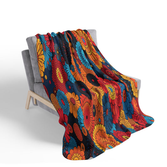 A soft gray armchair is draped with an African-inspired fleece blanket featuring a bold floral pattern in red, yellow, blue, and orange. The vivid flowers pop against the dark sherpa-backed fleece, creating a lively contrast in this cozy home decor setting.