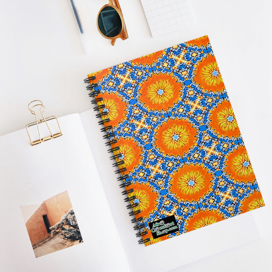 A culturally inspired notebook with a bright orange, blue, and yellow geometric floral pattern cover sits on a white table. Nearby are an open book with a photo, gold binder clip, brown sunglasses, and a white eraser in this minimalistic scene.