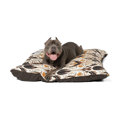 A gray dog with cropped ears and a pink tongue lies on a large, African-inspired patterned dog bed featuring brown, white, and orange floral designs. The dog appears relaxed and happy, facing forward with its mouth open and tongue out.