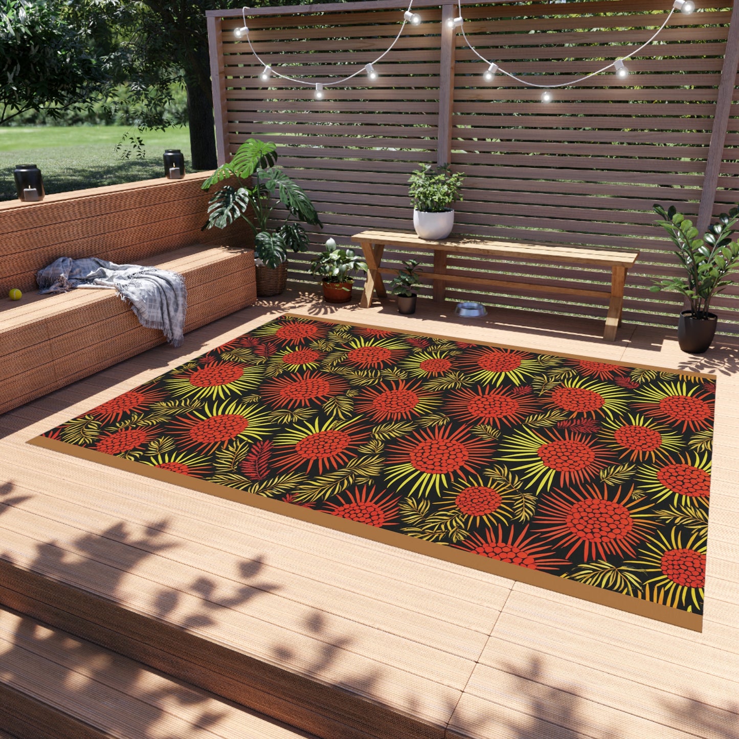 A cozy outdoor patio with wood decking, a bench, a cushioned sectional, potted plants, and string lights. An African-inspired rug with red and yellow tropical leaf patterns is centered on the deck, adding a bold accent to the relaxed setting.