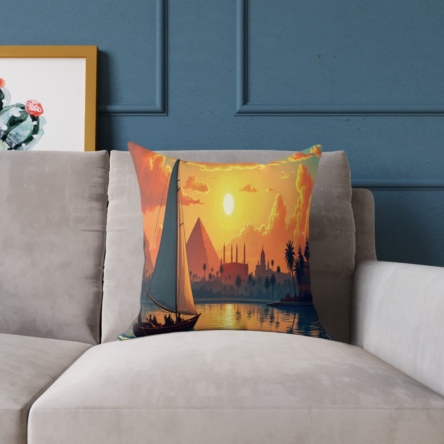 A colorful accent pillow with a vivid sailboat print, pyramids, domes, and palm trees rests on a light gray couch. Perfect as vibrant home decor, it pops against a blue wall with a framed cactus artwork partially visible on the left.