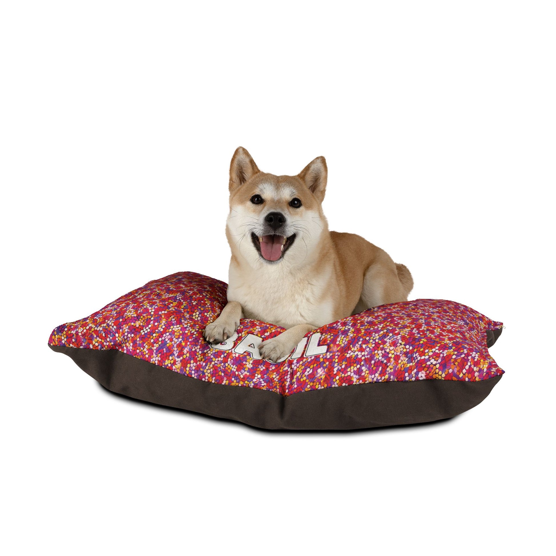 A Shiba Inu with tan and white fur lies happily on a large, colorful dog bed featuring durable construction and a red, pink, and white patterned top with brown sides. The dog faces forward, mouth open, tongue out, against a plain white background.