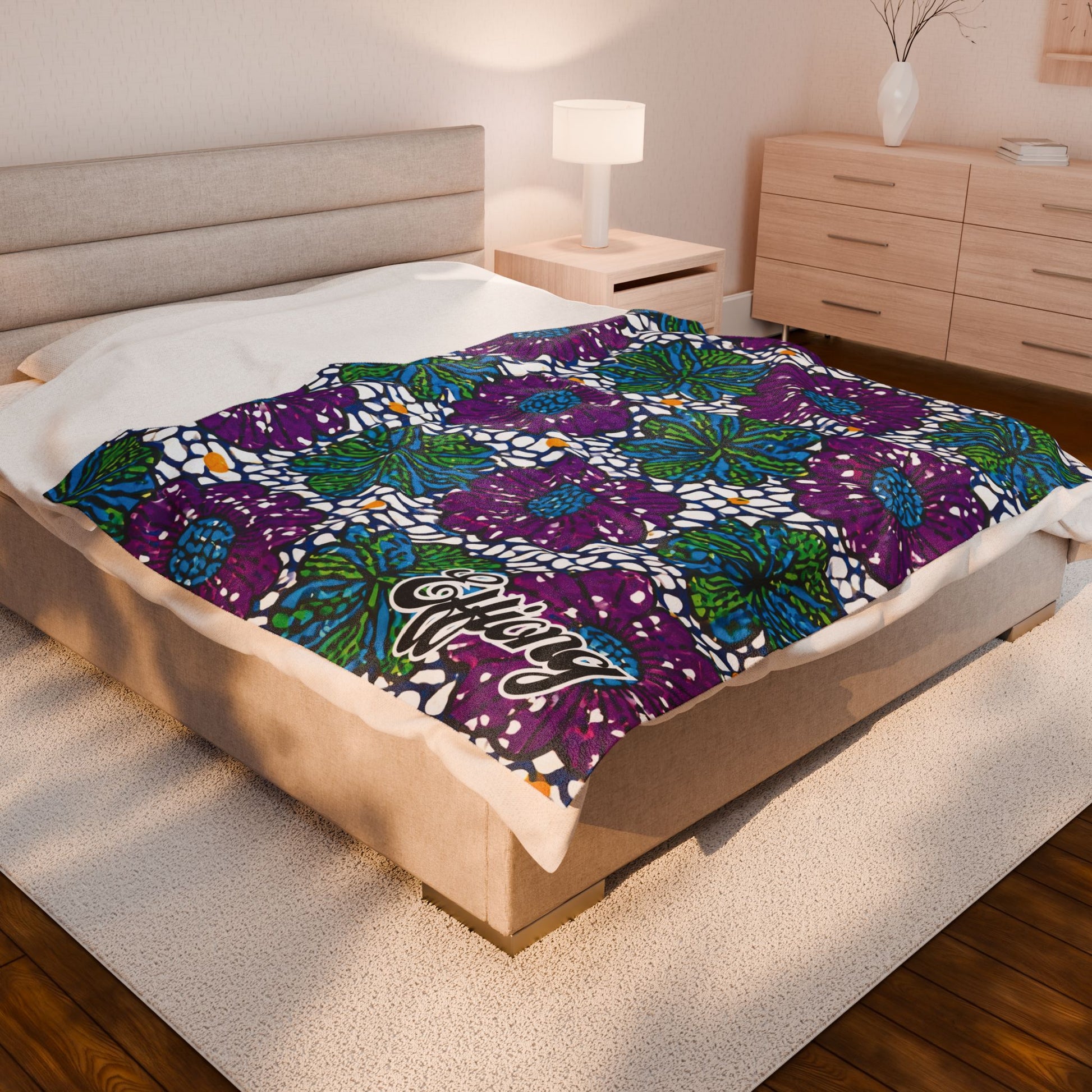 A cozy bedroom with a beige bed frame and nightstand. The bed is covered with an African-inspired personalized blanket, featuring bold geometric and floral patterns in purple, green, blue, and white script text. Warm decor and wooden floors complete the space.