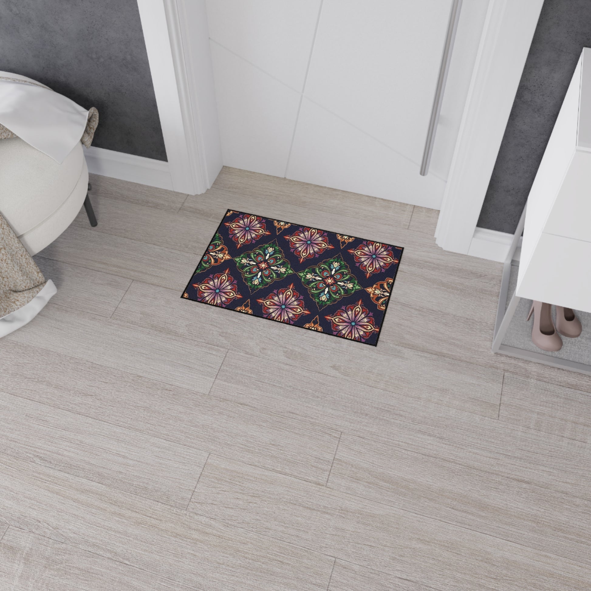 A small rectangular, non-slip doormat with colorful, intricate floral and geometric patterns sits on light wood flooring in a modern entryway. The door mat has a dark background, white walls, a shoe rack, and a round cushioned seat nearby.