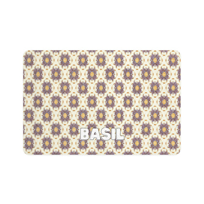 A rectangular mat with a repeating pattern of African-inspired, stylized purple and yellow flowers on a cream background. The word BASIL is written in bold, white, uppercase letters near the bottom left corner.
