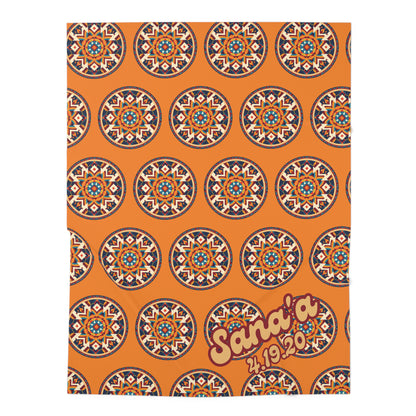 An orange baby swaddle blanket features a repeating pattern of circular geometric mandala designs in blue, white, and red. In the lower right corner, bold yellow text with a red outline reads “Sana’a 4.19.20.”, perfect for African-inspired decor.
