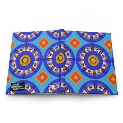 An open hardcover journal notebook displays a vibrant geometric pattern with blue, orange, and yellow circular starburst shapes and orange diamond motifs on a light blue background. The bottom left cover features a black label with white and yellow text and a QR code nearby.