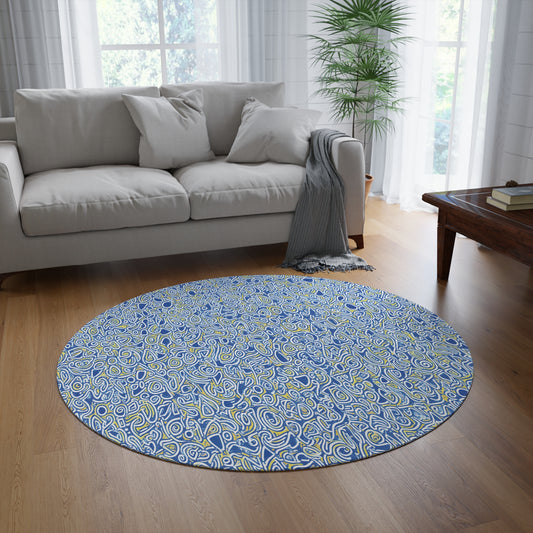 A round, African-inspired rug with an abstract blue, white, and yellow swirling pattern lies on a wooden floor in a bright living room. A light gray sofa with white pillows and a draped gray blanket sits nearby, framed by sheer curtains and a green potted plant.