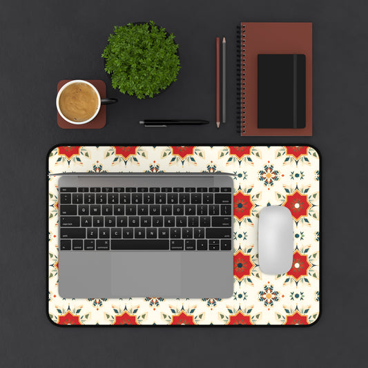 A top-down view of a workspace featuring a laptop and mouse on a colorful neoprene desk mat with floral patterns, a cup of coffee, small green plant, notebook, closed red folder, pen, and pencil—all arranged on a dark surface.