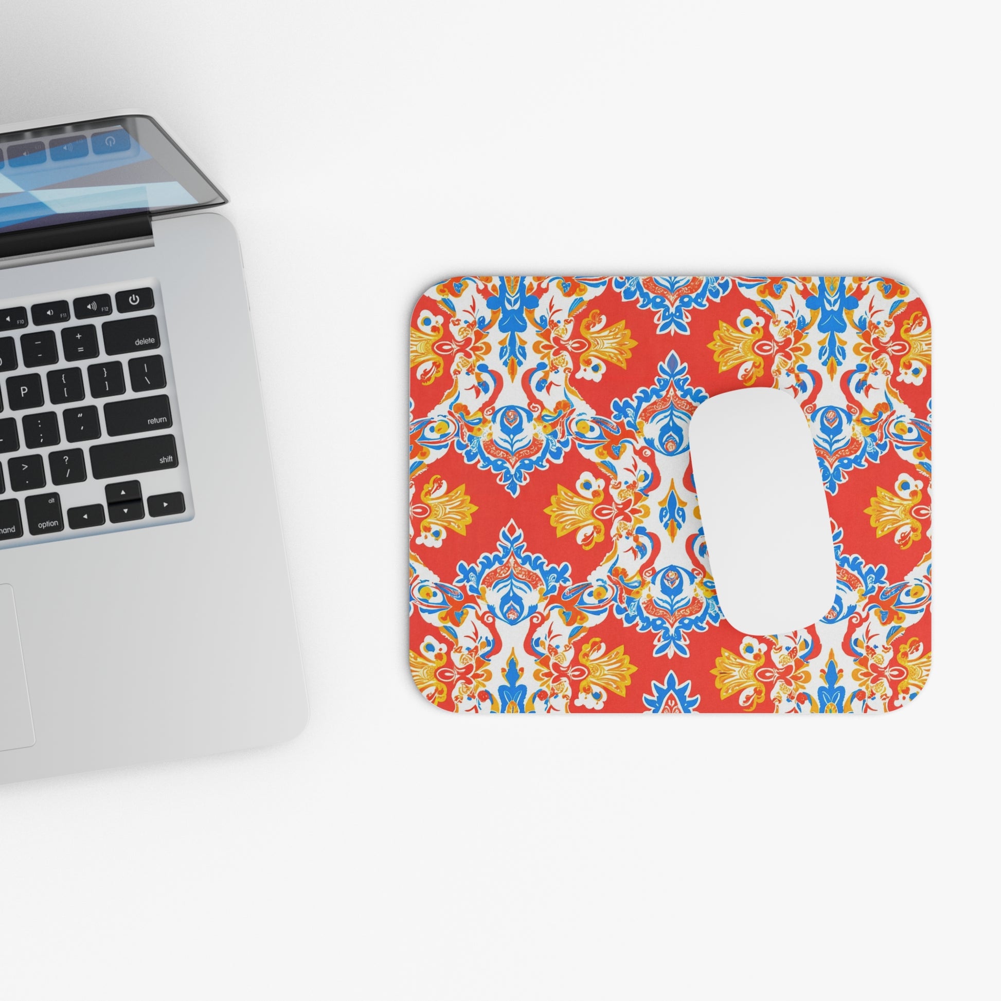 A silver laptop sits on a white surface next to a creative desk accessory: an African-inspired mouse pad with a symmetrical floral pattern in red, blue, and yellow. A white wireless mouse rests on its non-slip rubber base. The scene is brightly lit.