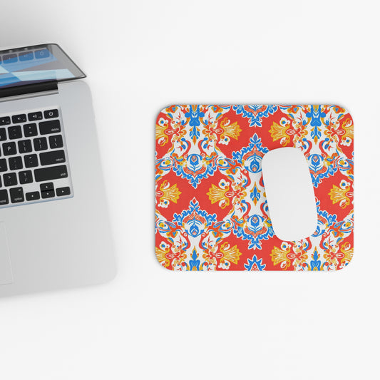 A silver laptop sits on a white surface next to a creative desk accessory: an African-inspired mouse pad with a symmetrical floral pattern in red, blue, and yellow. A white wireless mouse rests on its non-slip rubber base. The scene is brightly lit.