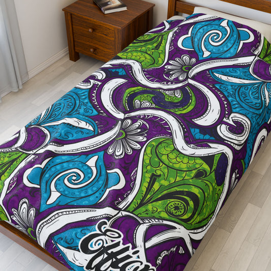 A wooden bed with a colorful, African-inspired blanket featuring abstract, swirling paisley patterns in blue, green, purple, black, and white. The bed sits beside a wooden nightstand holding a book, against light-colored floors and walls.