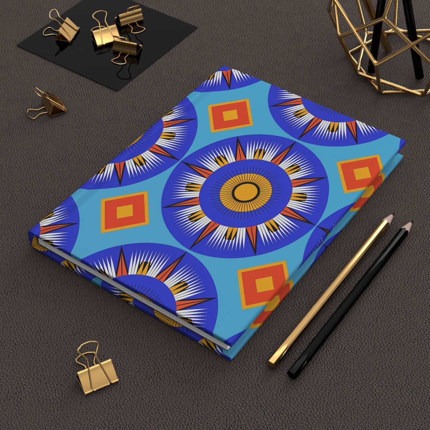 A closed hardcover journal notebook with a vibrant blue, orange, and yellow geometric sunburst pattern, reminiscent of an African-inspired journal, lies on a brown textured surface beside black and gold pencils, gold binder clips, and modern geometric decor.