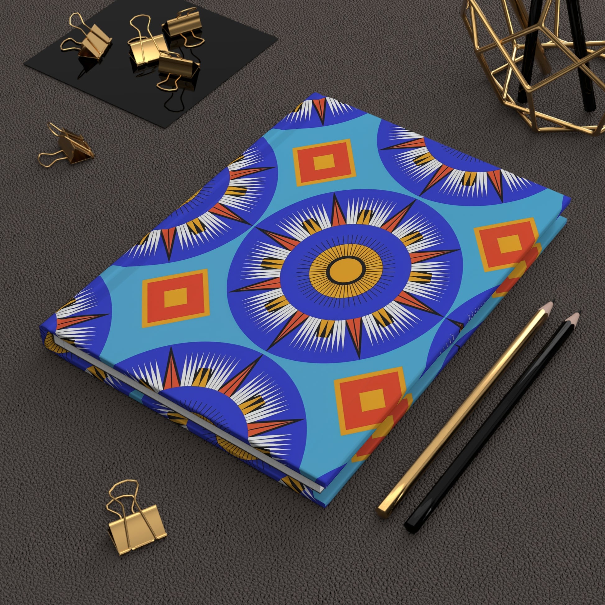 A closed hardcover journal notebook with a vibrant blue, orange, and yellow geometric sunburst pattern, reminiscent of an African-inspired journal, lies on a brown textured surface beside black and gold pencils, gold binder clips, and modern geometric decor.