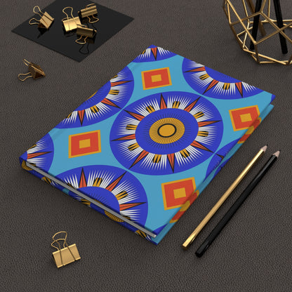A closed hardcover journal notebook with a vibrant blue, orange, and yellow geometric sunburst pattern, reminiscent of an African-inspired journal, lies on a brown textured surface beside black and gold pencils, gold binder clips, and modern geometric decor.