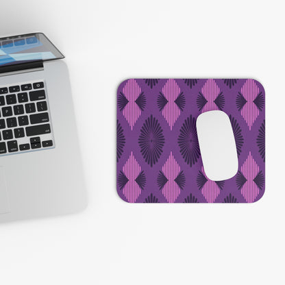 A silver laptop sits on a white surface next to a creative desk accessory: an African-inspired mouse pad with alternating dark purple and magenta diamond shapes, radiating lines, and a non-slip rubber base. A white computer mouse rests on top.