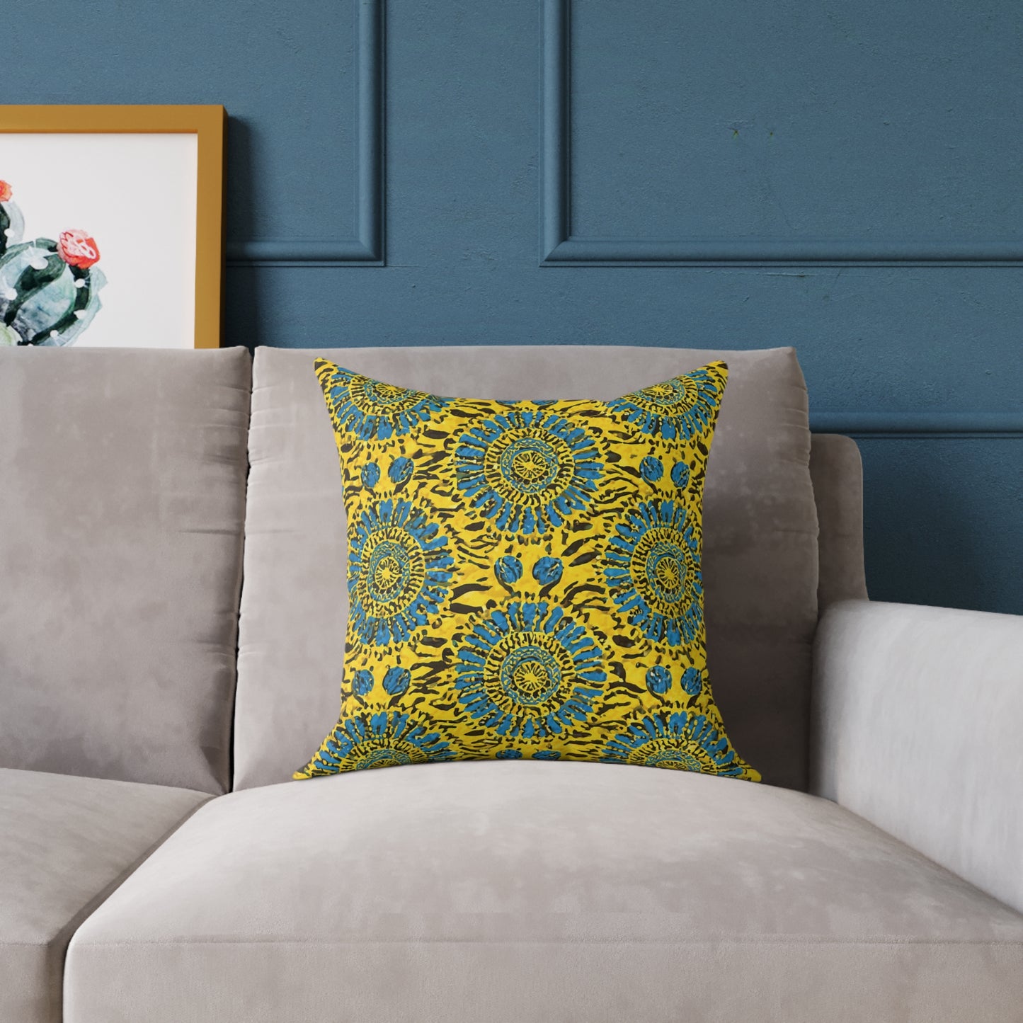 A colorful accent pillow featuring a blue and green floral mandala pattern rests on a light gray couch. Behind the couch is a dark blue paneled wall, with part of a framed cactus illustration visible to the left, adding vibrant home decor flair.