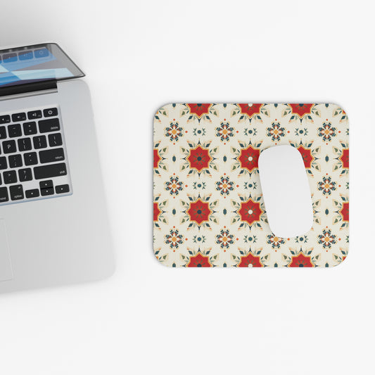 A silver laptop sits on a white surface next to a vibrant desk accessory—an African-inspired mouse pad with a non-slip rubber base. The pad features repeating geometric patterns with red star shapes, blue accents, and floral motifs on cream. A white mouse rests on it.