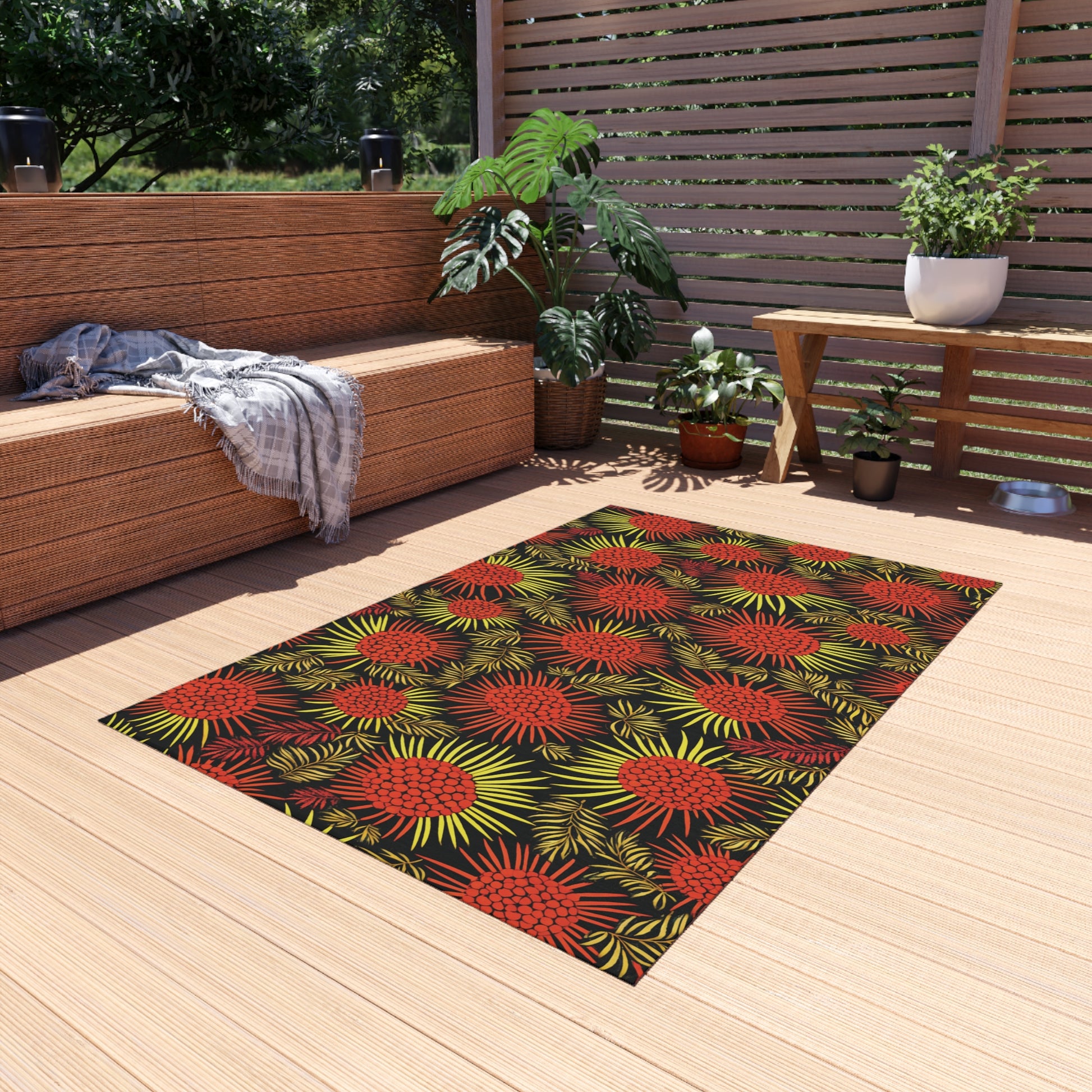 A modern patio with wooden decking features an African-inspired rug with a bold red and yellow floral pattern. Wicker furniture with gray cushions, a throw blanket, potted plants, and a slatted wooden privacy screen complete the relaxed outdoor setting.