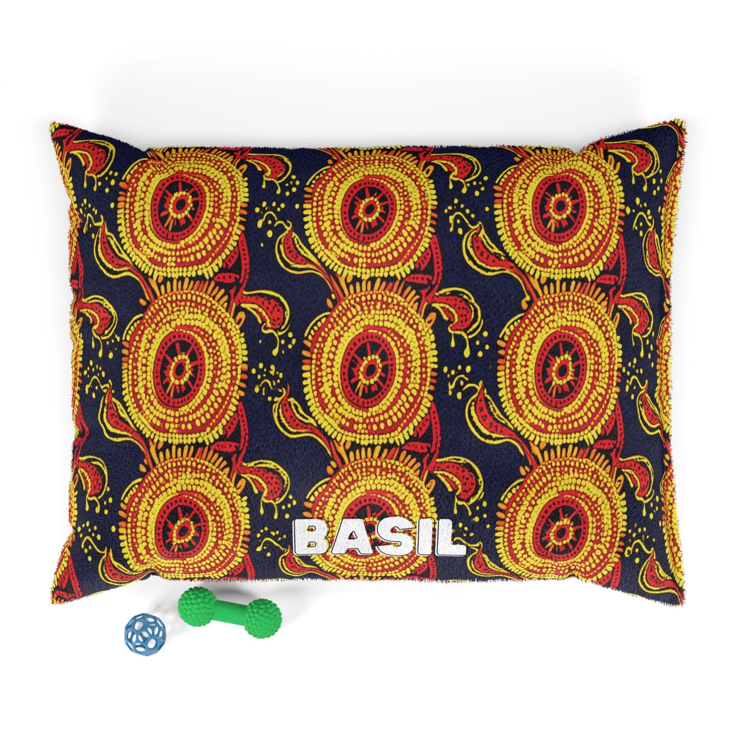 A rectangular pillow with a vibrant African-inspired red, yellow, and black circular pattern. The word “BASIL” in large white letters is on the front. Below the pillow are two pet toys: a green bone-shaped toy and a blue ball. The background is white.