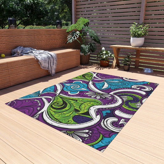 A modern patio with light wood decking features a weather-resistant outdoor rug in a colorful, abstract pattern of green, purple, blue, and white. Wicker furniture with neutral cushions, potted plants, and a wooden privacy screen complete the inviting space.