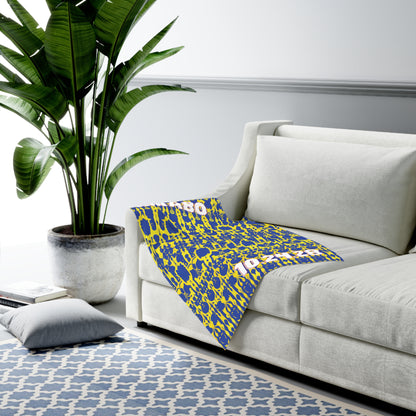A modern living room with a light gray sofa, an African-inspired decor touch from a blue and yellow patterned blanket draped over the arm, a gray potted plant, blue and white geometric rug, and a gray floor cushion. The room has soft natural lighting.