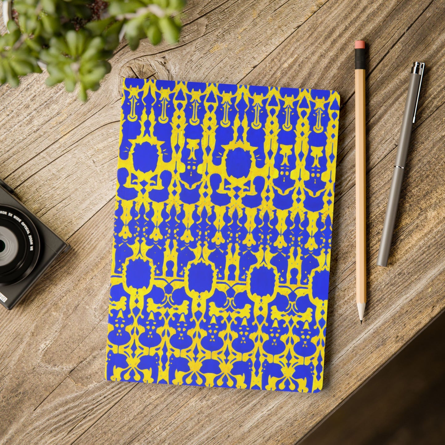 A bold yellow and blue African-inspired journal lies on a rustic wooden table next to a pencil, a pen, a camera, and a potted plant in the corner, creating a creative and organized workspace vibe perfect for daily journaling.