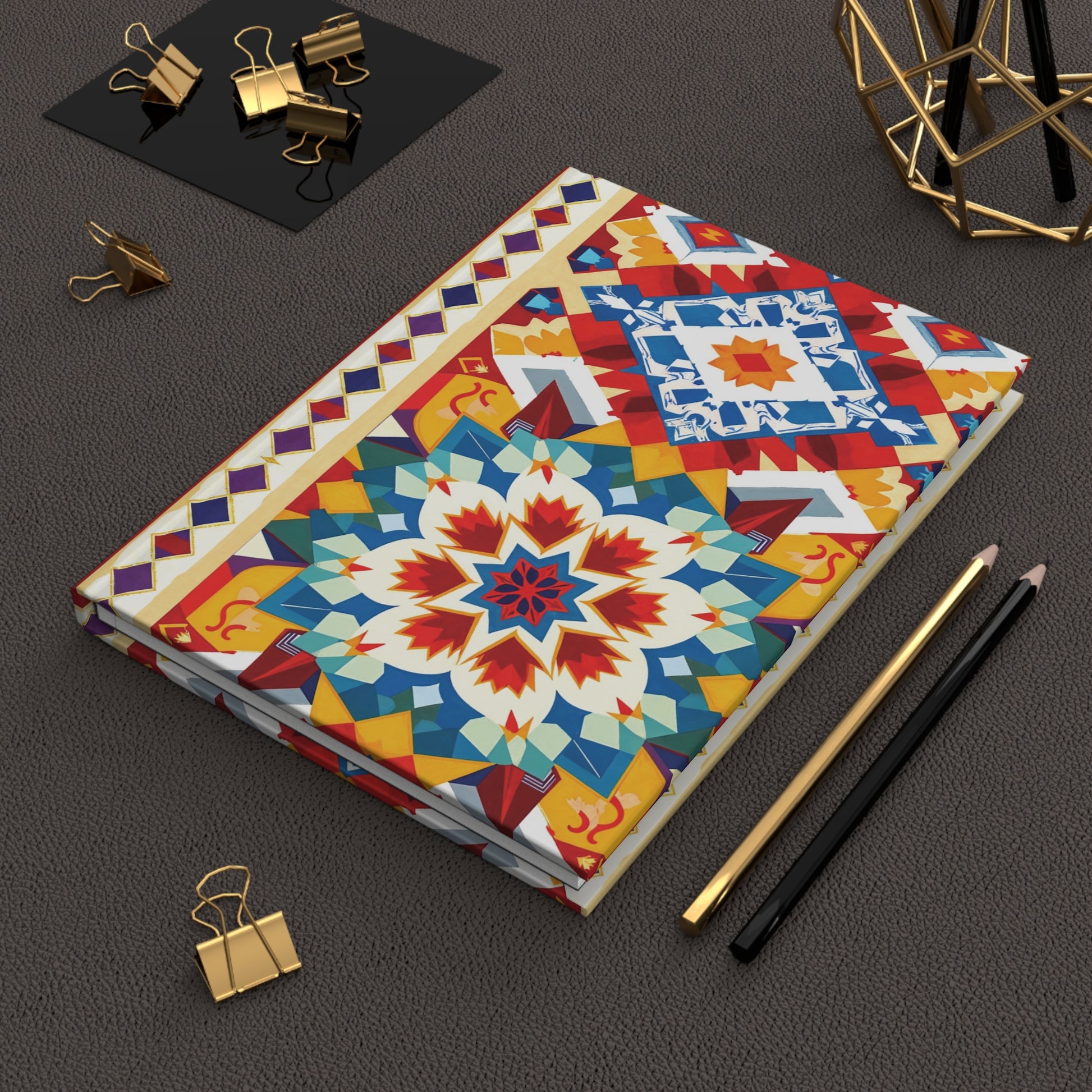 A closed hardcover journal notebook with a vibrant, African-inspired design and floral pattern rests on a textured brown surface. Nearby are black and gold pencils, gold binder clips, and a decorative geometric metal object—all neatly arranged.