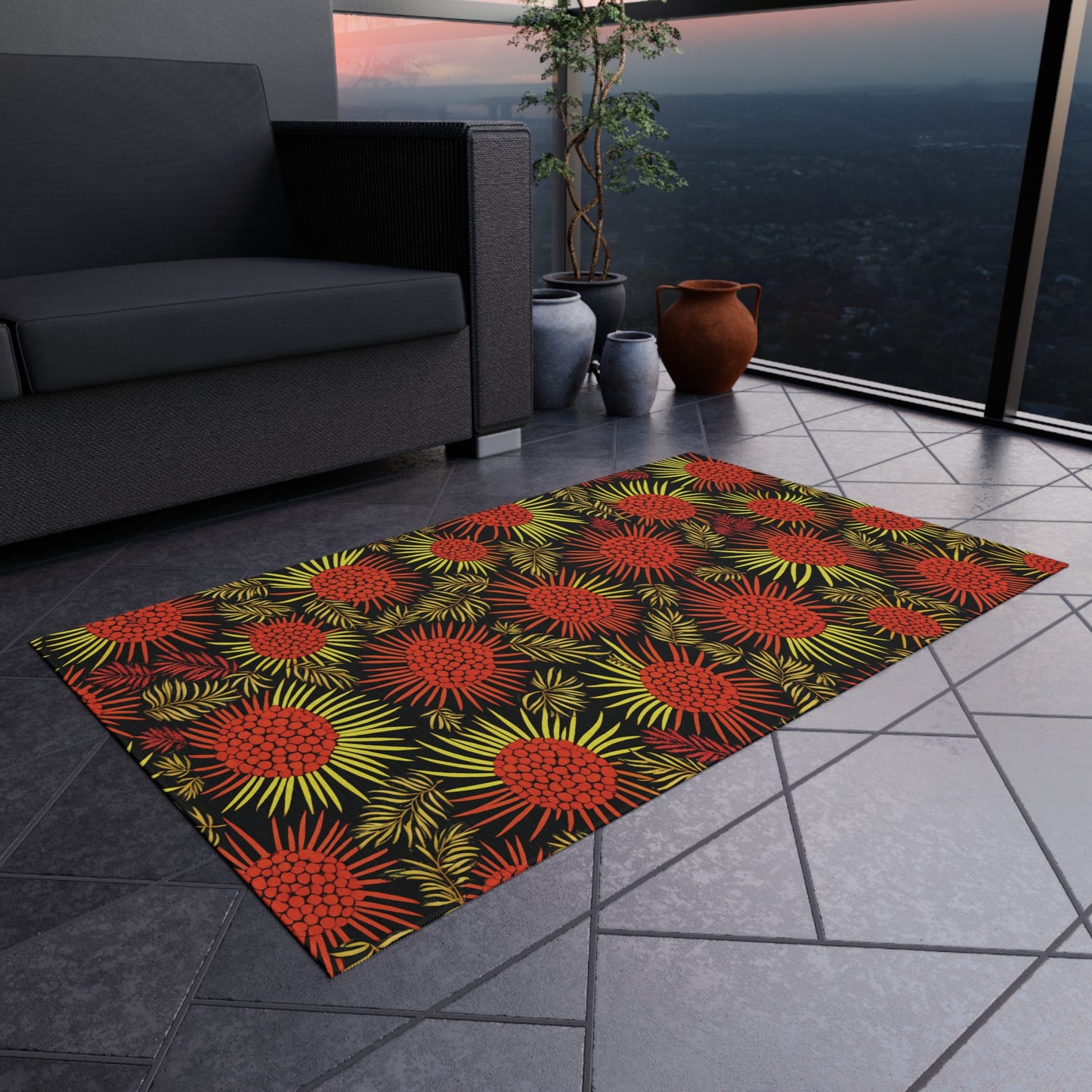 A rectangular rug with bold red and yellow African-inspired tropical floral patterns lies on a gray-tiled balcony floor. Nearby are a dark sofa, a leafy plant in a black pot, a brown clay pot, and a railing overlooking the city at dusk.
