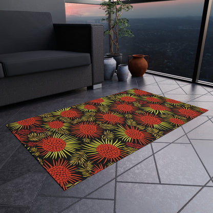 A rectangular rug with bold red and yellow African-inspired tropical floral patterns lies on a gray-tiled balcony floor. Nearby are a dark sofa, a leafy plant in a black pot, a brown clay pot, and a railing overlooking the city at dusk.