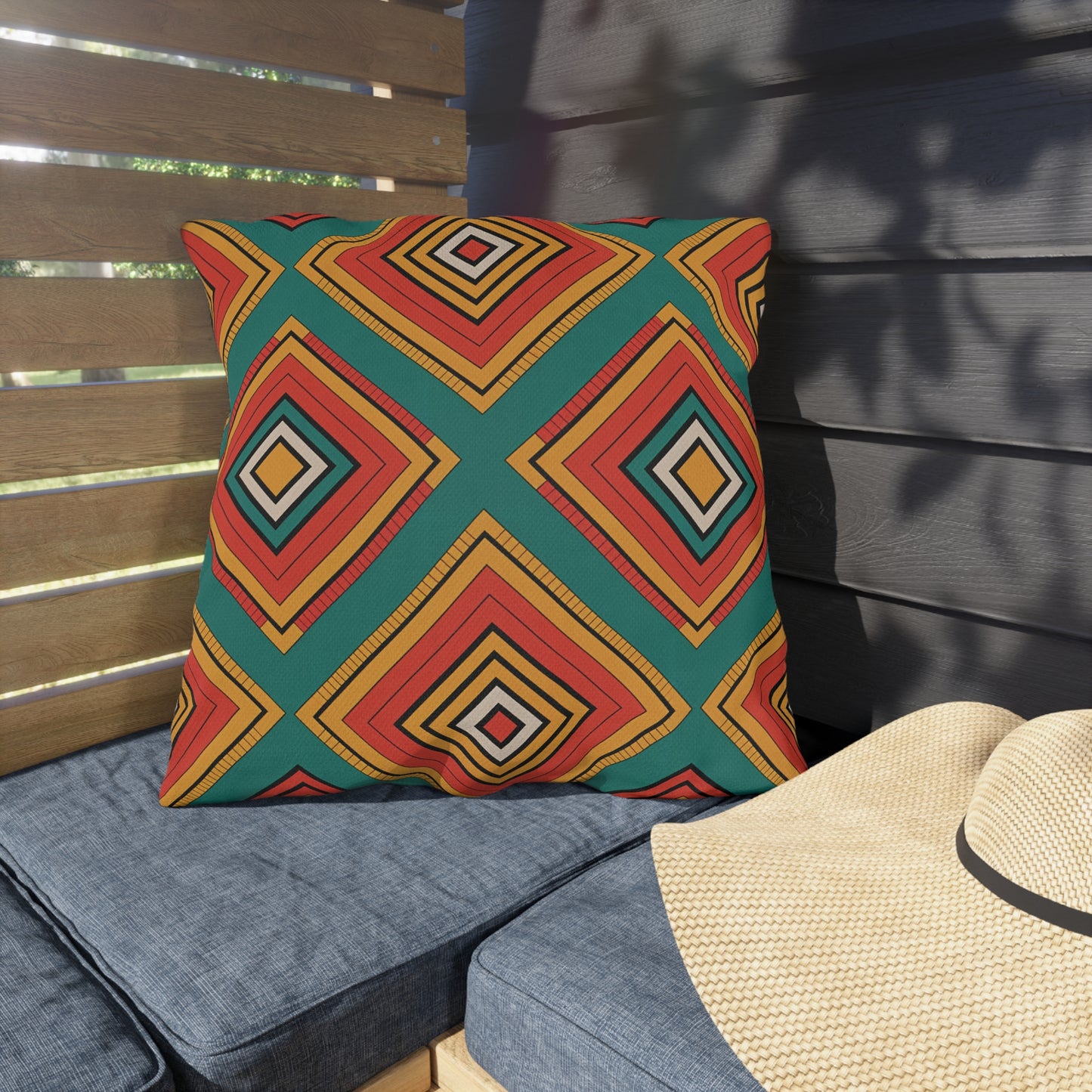 A square outdoor pillow with a bold geometric pattern in red, yellow, black, and green sits on a blue cushion in an outdoor seating area. Made from weatherproof materials, it adds style while sunlight dances across the wooden wall behind it.