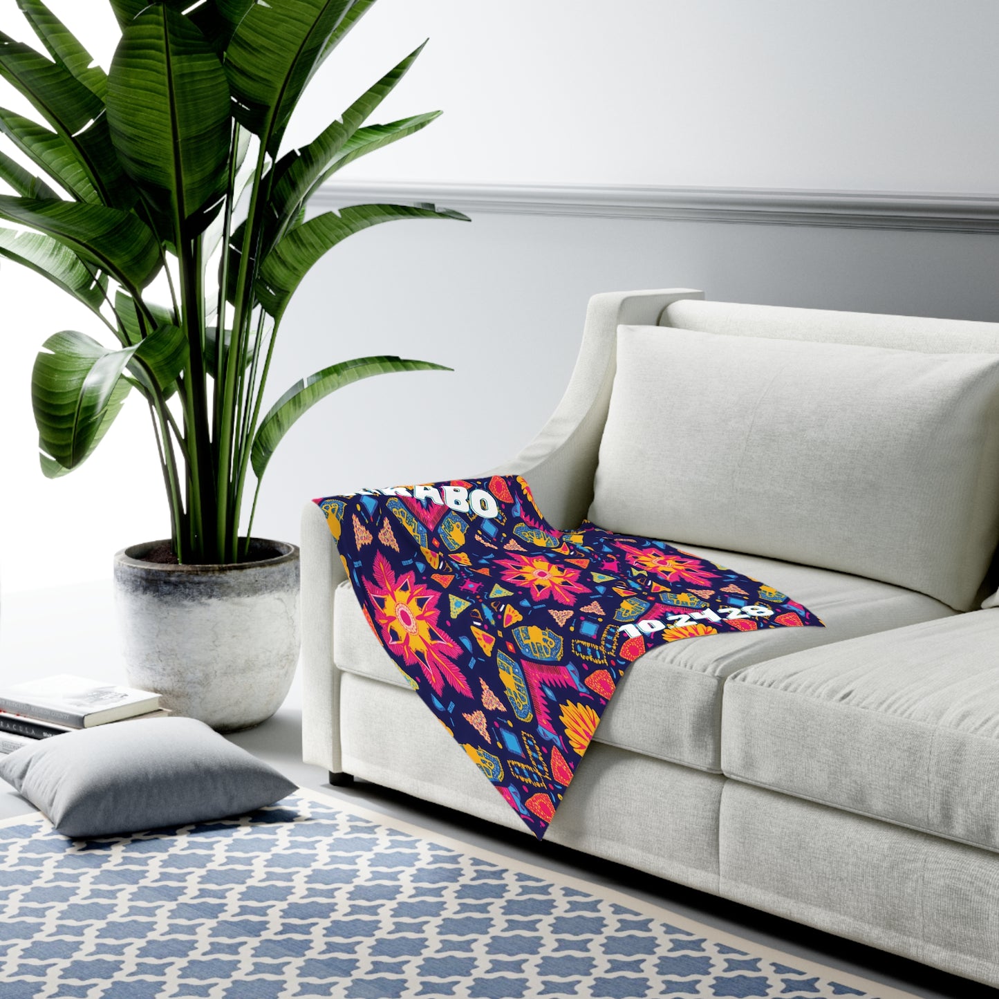A modern living room with African-inspired decor features a large potted plant, a white L-shaped sofa, and a colorful blanket with vibrant floral and geometric patterns. A blue and white patterned rug and gray floor pillow rest on the light gray walls.