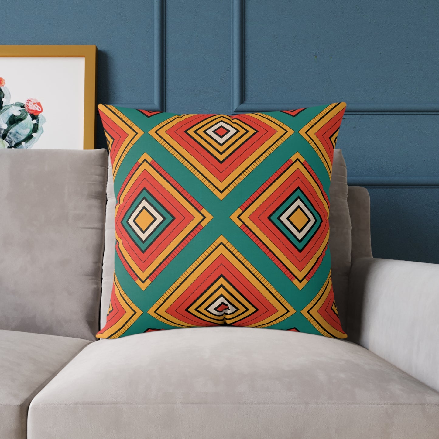 A vibrant throw pillow with a bold geometric diamond pattern in red, yellow, orange, and black on a teal background sits on a light gray couch. The couch is in front of a textured blue wall, with a framed cactus print partially visible on the left.