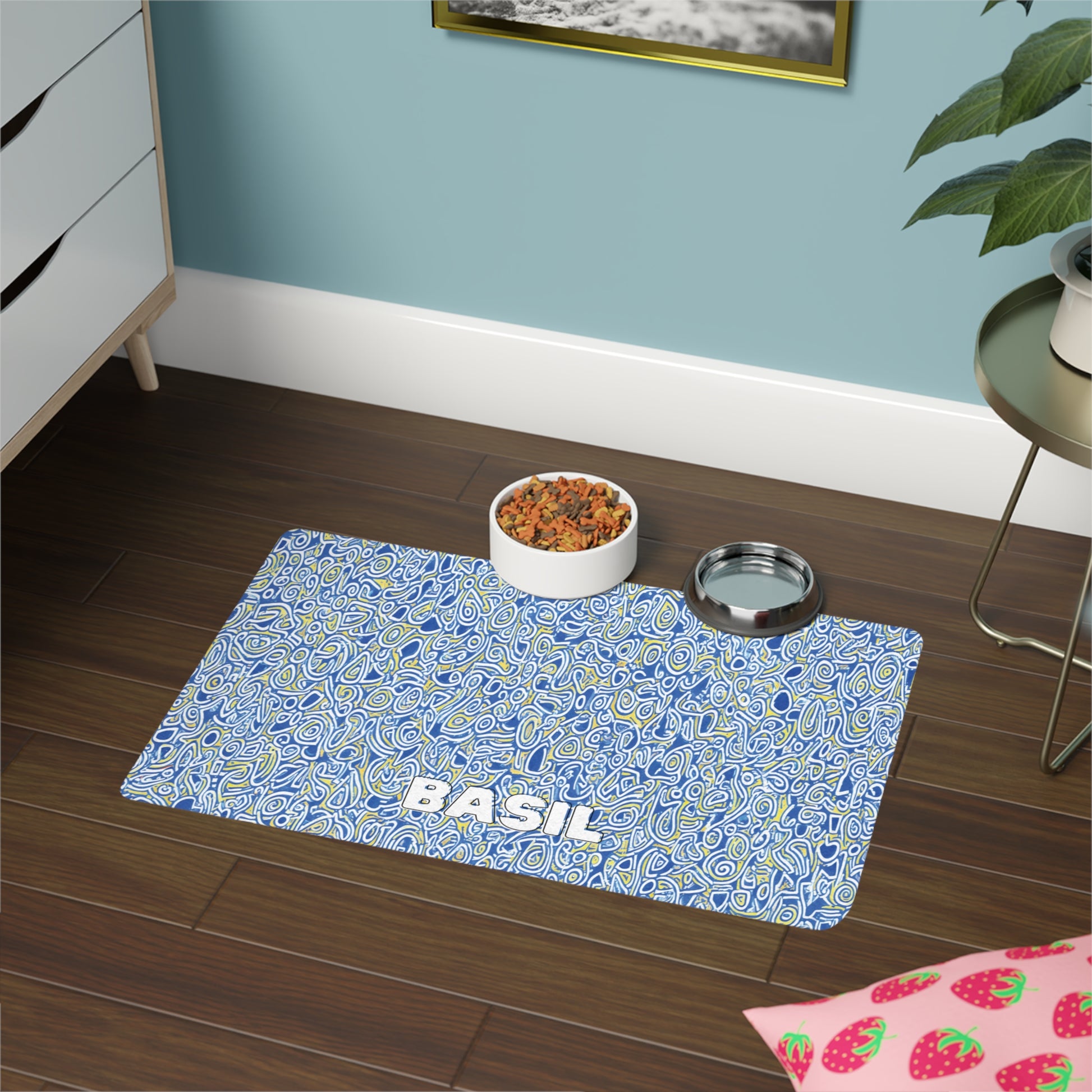 A blue, African-inspired patterned pet placemat with the name BASIL in white letters sits on a dark wood floor. On the mat are a bowl of kibble and a metal water bowl. Nearby are a green plant, white dresser, and strawberry-patterned mat.
